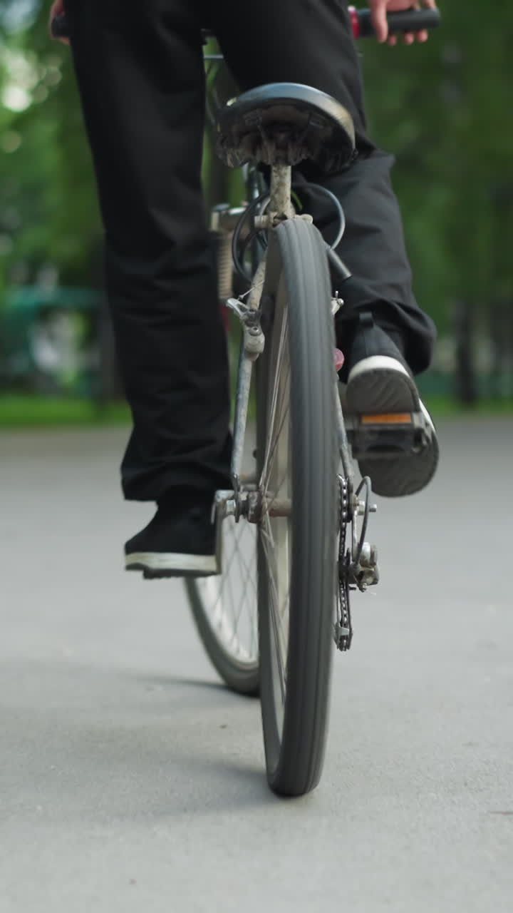 vista trasera de una persona en zapatillas de deporte de pie mientras monta una bicicleta en una carretera pavimentada de manera irregular, capturada desde un ángulo bajo, en el fondo, otra persona sentada en un banco