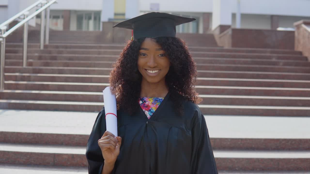 una joven afroamericana graduada de pie frente a la cámara con un diploma y libros en las manos. la estudiante está de pie en las escaleras afuera.