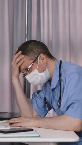 young doctor, in protective mask, writes with ballpoint pen, fills medical card, sits in his office at table, holds his hand on uncut hair, fatigue, impotence, tired