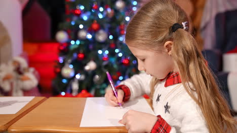 child writing a letter to santa claus. cute little girl sitting at the table in christmas decorated room and drawing on paper. winter holidays. christmas concept.