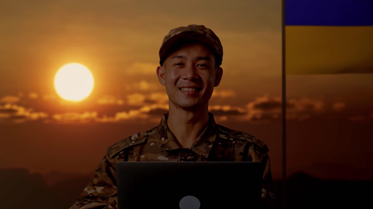 Soldato asiatico che usa un portatile al tramonto con la bandiera ucraina sullo sfondo