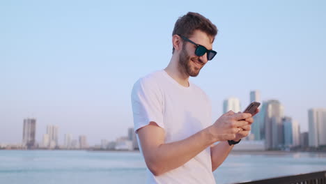 Attraktiver-Und-Gutaussehender-Junger-Mann-Mit-Schönem-Lächeln-Und-Hipster-Look-Der-Millennial-Generation-Z-Scrollt-Durch-Den-Social-Media-Feed-Auf-Dem-Smartphone,-Kichert-Und-Lächelt-Bei-Den-Nachrichten-In-Den-Anwendungen.