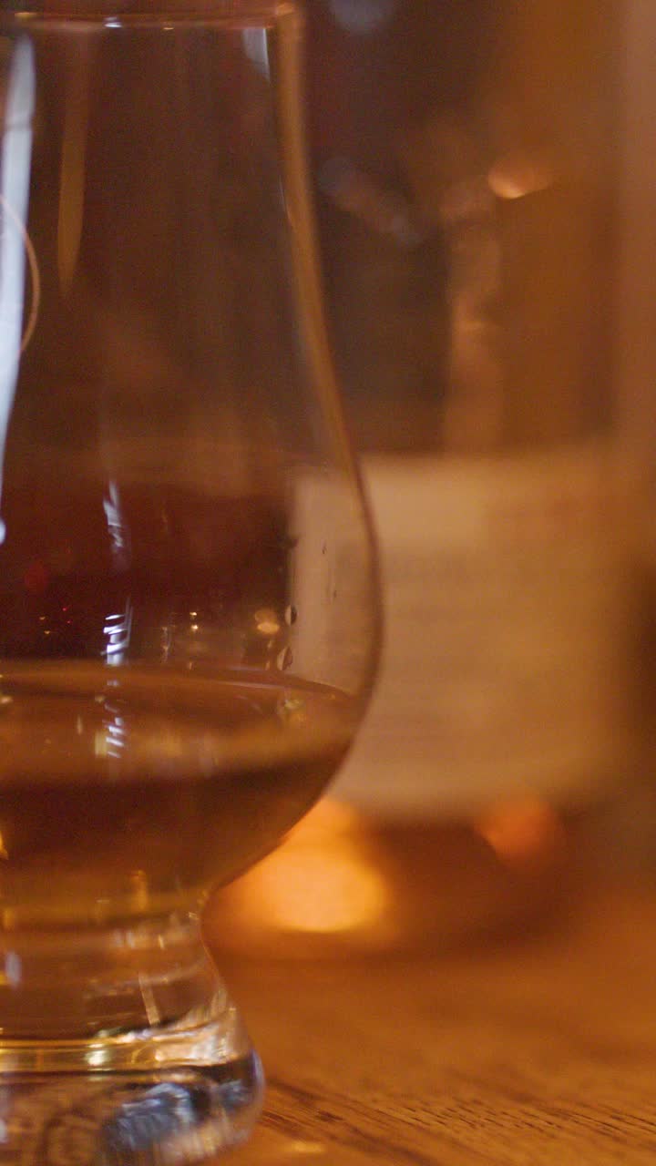Una mano posa un bicchiere di whisky su un bancone di legno; l'illuminazione calda e le bottiglie sfocate evocano un pub accogliente.
