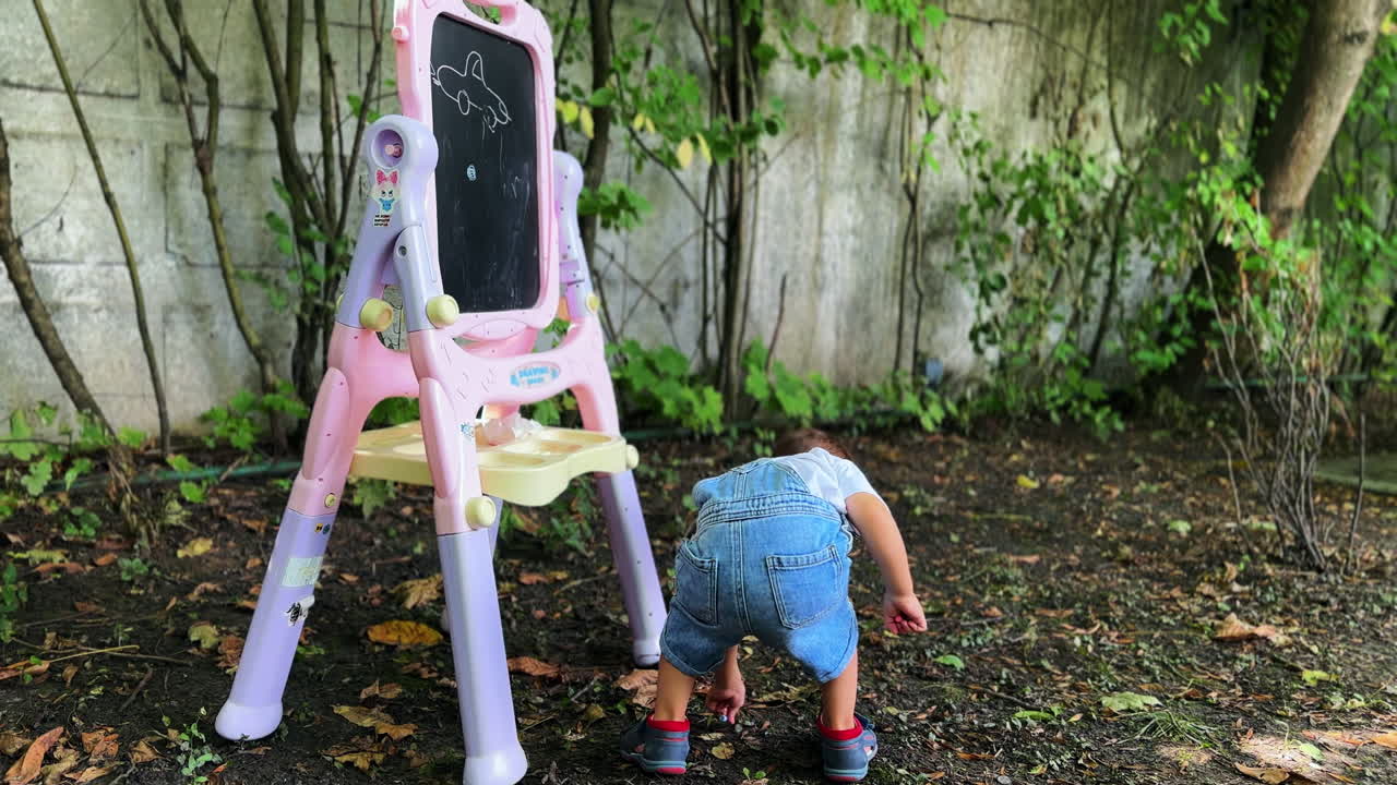 Little toddler boy staying outdoors near the blackboard. Kid drops the chalk and picks it up.