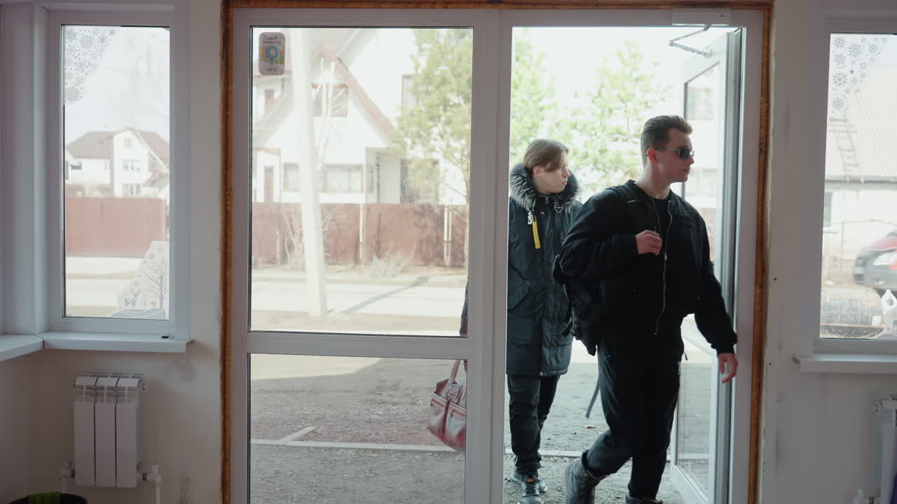 Two guys in dark jackets walked into glass entrance of building, one holding leather bag, other adjusting sunglasses, seen through door from inside room with radiators and bucket, urban moment in daylight