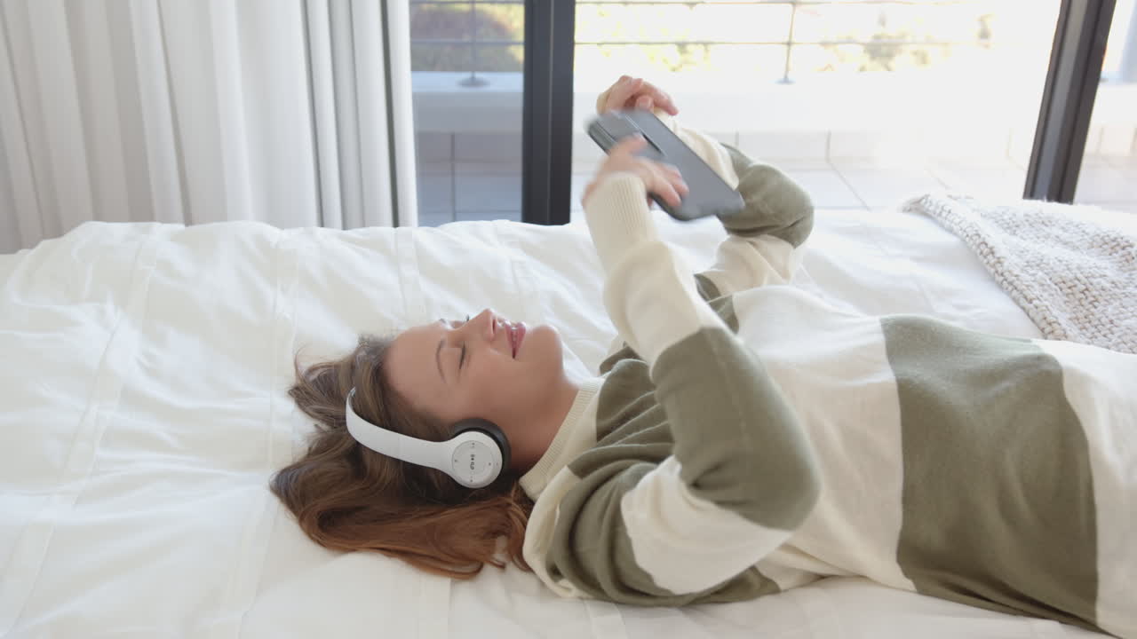 Relaxing on bed, woman listening to music with headphones and holding smartphone