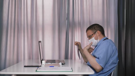 Arzt-In-Maske-Hält-Konferenz-über-Tablet-Und-Laptop-Am-Tisch