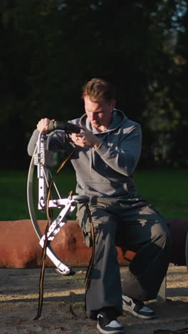 person in gray hoodie and dark pants focused on adjusting stilts in sunny outdoor setting surrounded by lush greenery. concentrated expression shows attention to detail, with natural light