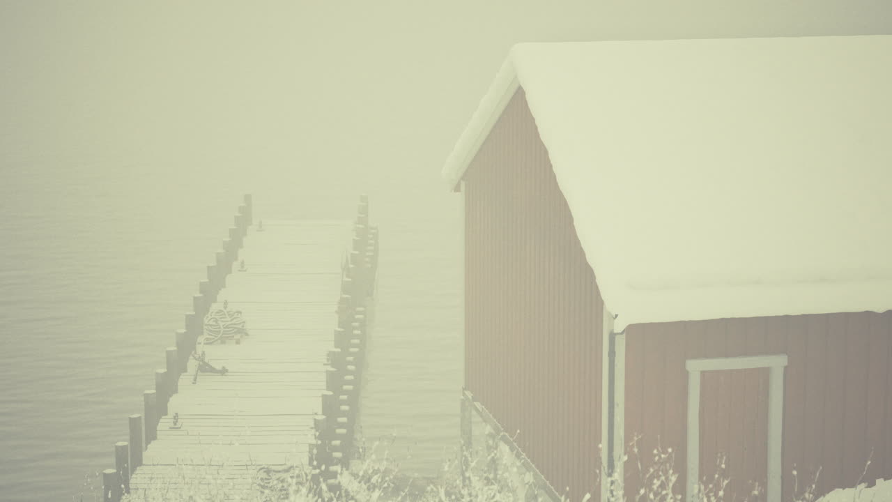 Snowy winter mist envelops wooden cabin by frozen lake pier in serene landscape