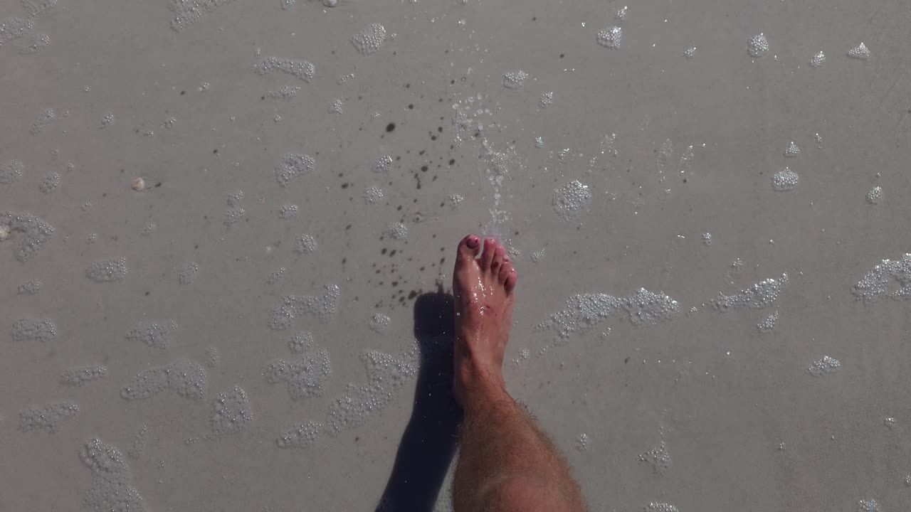A barefoot person slowly steps across smooth wet sand as gentle ocean foam washes the shore. The slow-motion captures each movement with relaxing detail.