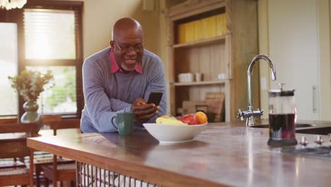 African-american-senior-man-leaning-on-kitchen-counter-using-smartphone,-taking-off-glasses,-smiling