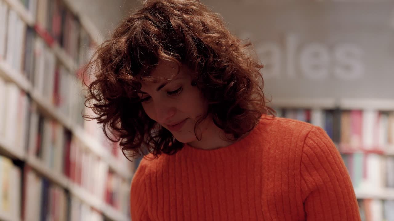Donna caucasica riccia concentrata che studia tra gli scaffali dei libri in biblioteca.