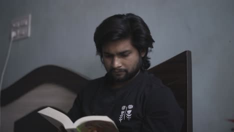un niño indio del sur de asia leyendo un libro en una habitación fría y oscura sentado en una cama