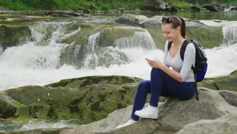 Una-Persona-Usa-Un-Teléfono-Inteligente-En-Una-Caminata-Sentada-Junto-A-Un-Hermoso-Río-De-Montaña