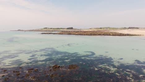 Reveal-drone-footage-of-golden-beach-with-crystal-clear-water,over-rocks-,-harbour-wall-and-second-beach-with-drying-boat-at-low-tide-in-Guernsey-on-bright-sunny-day