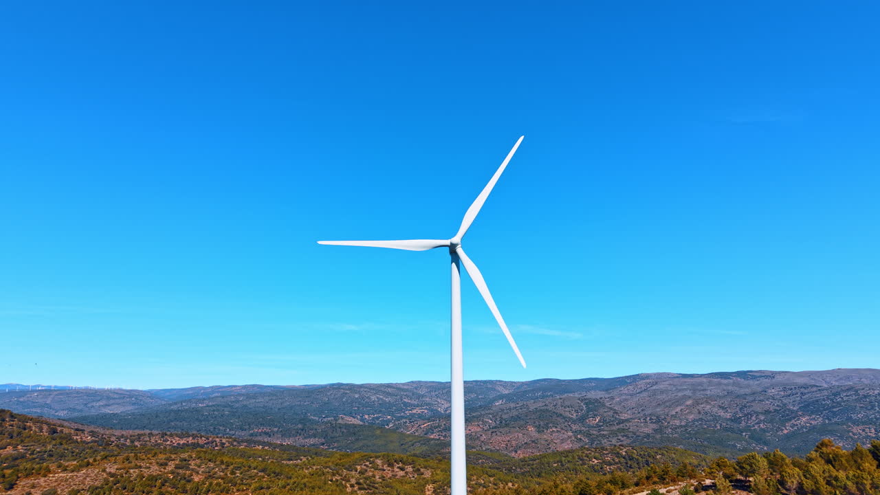 Madrid, Spagna, 8 agosto 2025: Primo piano di una singola turbina eolica contro un cielo azzurro limpido. Turbina eolica a tre pale centrata, torre bianca, sfondo di colline verdi, giornata di sole splendente, prospettiva dal basso.