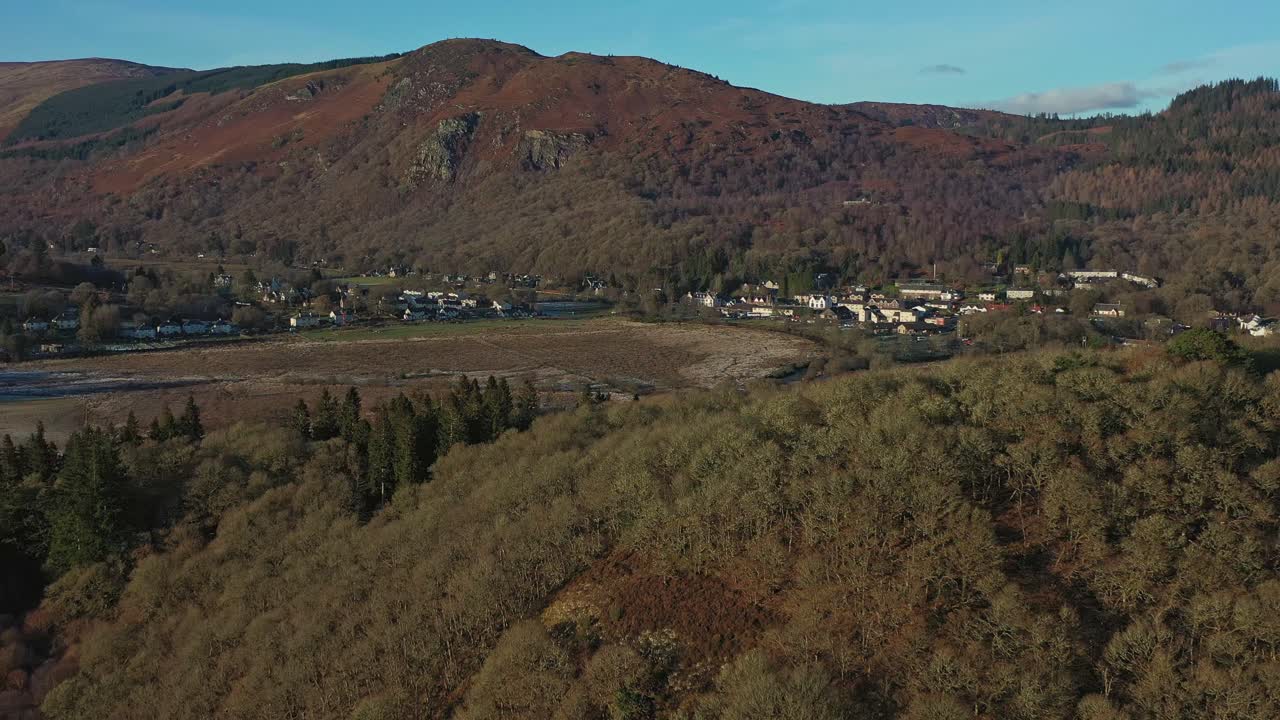 revelación aérea con una toma panorámica a través de la ladera del bosque en el pueblo de aberfoyle con craigmore en el fondo, escocia