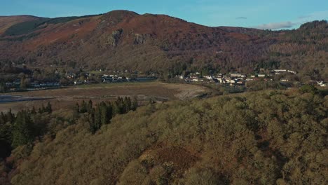 Revelación-Aérea-Con-Una-Toma-Panorámica-A-Través-De-La-Ladera-Del-Bosque-En-El-Pueblo-De-Aberfoyle-Con-Craigmore-Al-Fondo,-Escocia