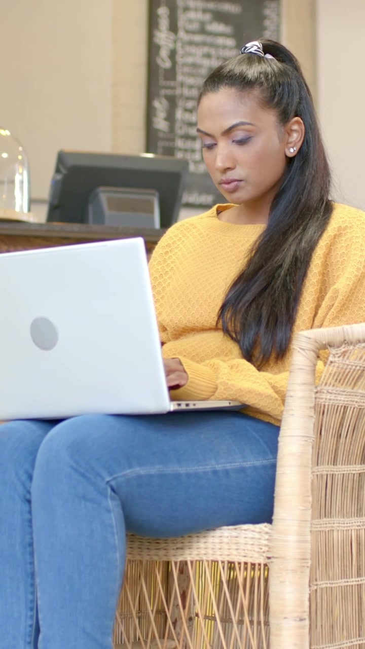 Vertical video: With laptop open, typing Indian woman in mustard knit working at cafe, white laptop