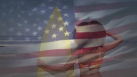 American-flag-waving-against-rear-view-of-woman-holding-surf-board-standing-on-the-beach