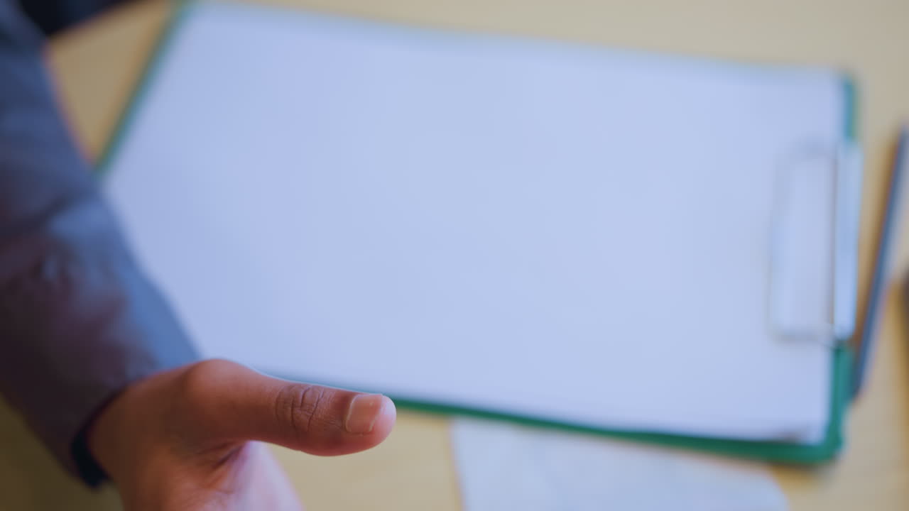 Close-up of man gently holding pen over paper with clipboard in background, suggesting moment of thought, pause, or preparation before writing in quiet workspace environment with soft lighting