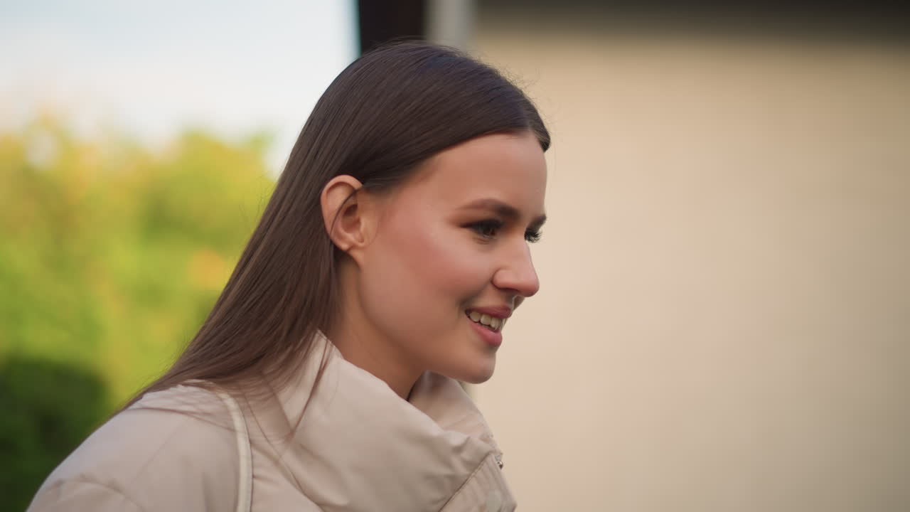Giovane donna all'aperto con cappotto beige che esprime un momento contemplativo da pendolare con un sorriso leggero e di profilo. Luce serale soffusa, sfondo verde sfocato, capelli lunghi lisci, trucco neutro