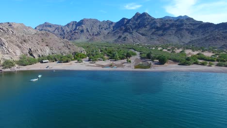 Forward-Flying-Aerial-Towards-Fishing-Village-on-Beach-Front-with-Flying-Birds-and-Dramatic-Mountains