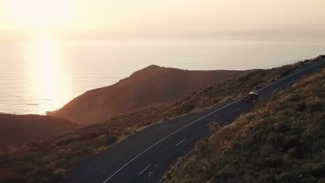 RED-TRUCK-DRIVING-AROUND-COASTAL-ROAD-AT-SUNSET-DRONE-FLYING-STRAIGHT