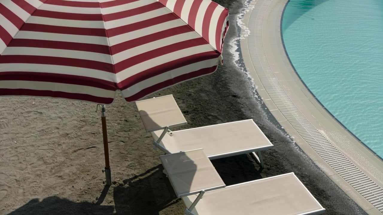 Telecamera in panoramica che mostra un ombrellone rosso e bianco con tre lettini bianchi a bordo piscina.
