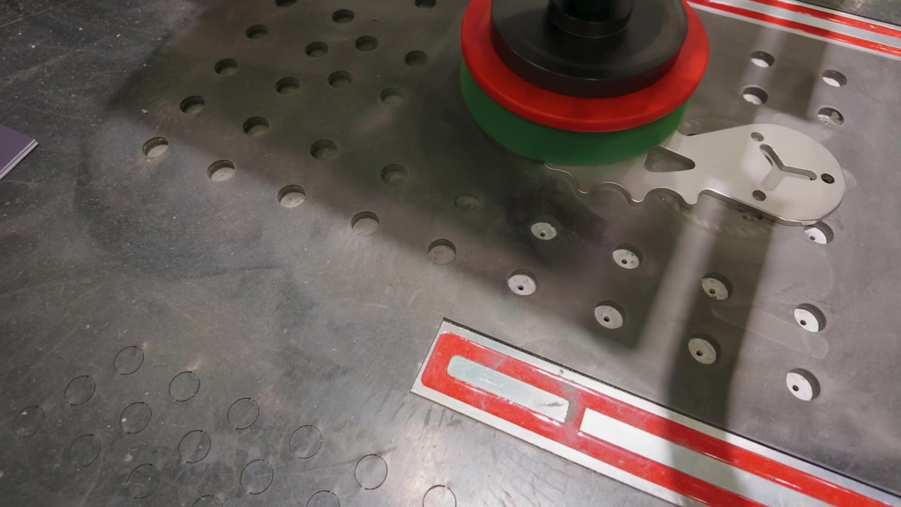 Deburring and linishing machine smooths sharp edges on a metal plate on a perforated workbench, showing abrasive head rotation and surface finishing in a workshop.