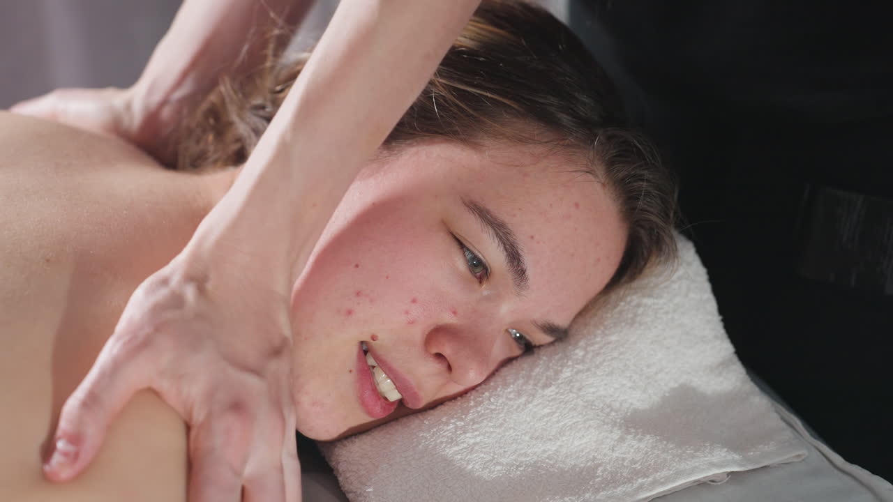 Close view of light skin blond woman lying peacefully on soft towel while receiving focused shoulder massage from therapist, showcasing gentle pressure, skin contact, and natural light reflections