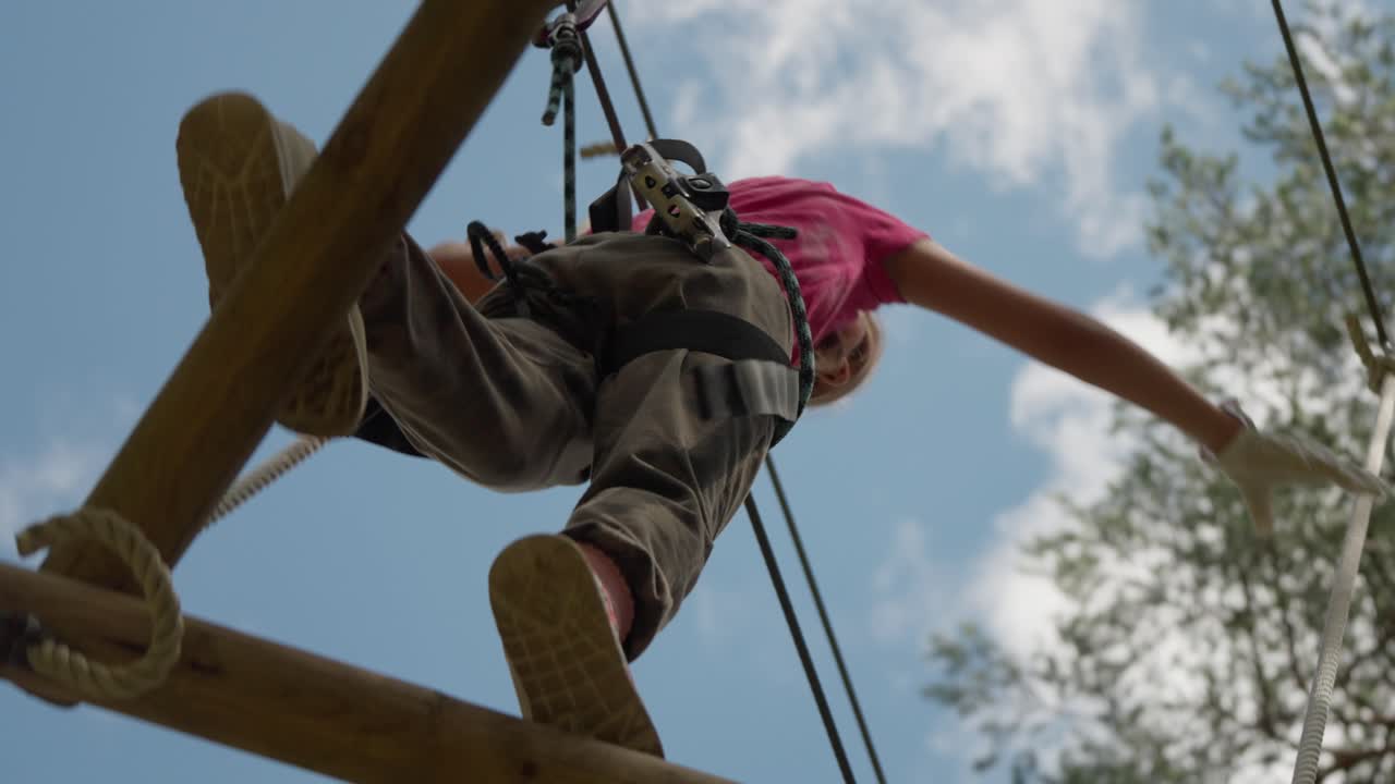 Person navigating high ropes course at adventure park in Stockholm. Individual wearing safety harness balances on wooden beam during summer outdoor activity. Low angle view against blue sky