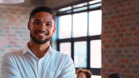 Portrait-Of-Smiling-Businessman-Standing-In-Busy-Office-With-Colleagues-Working-In-Background