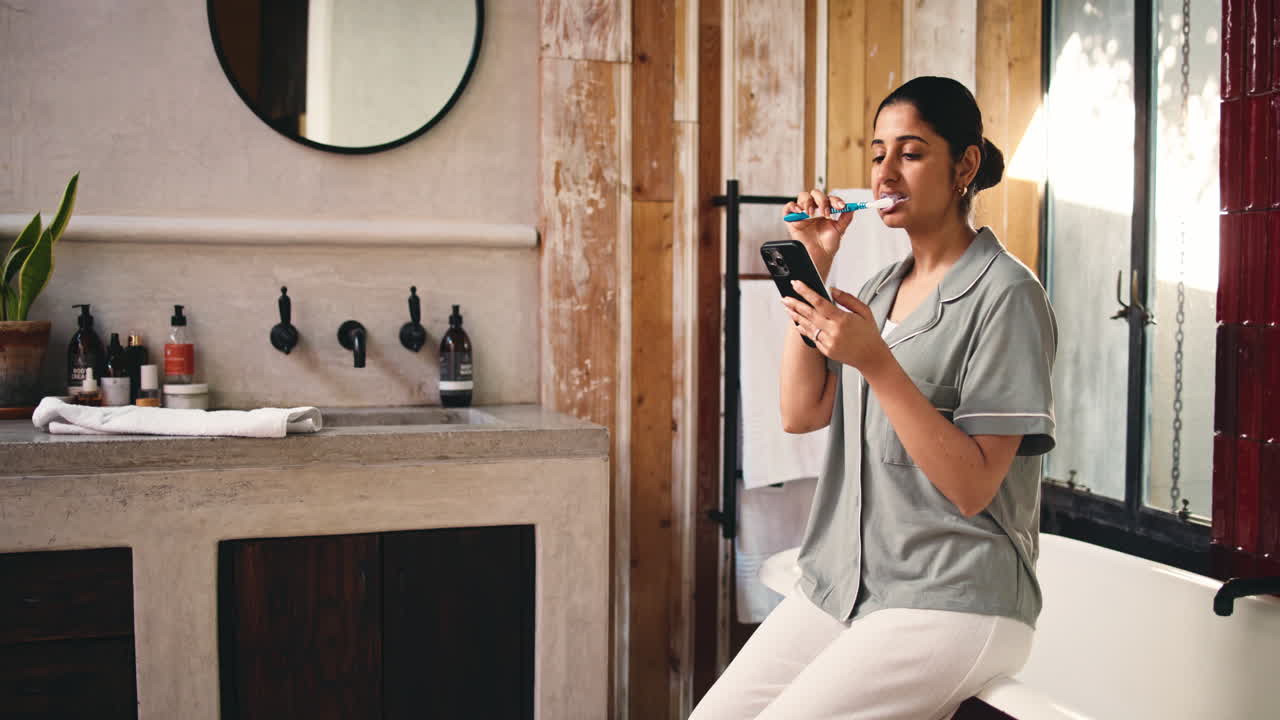 Giovane donna che si lava i denti e usa il telefono in un bagno moderno.