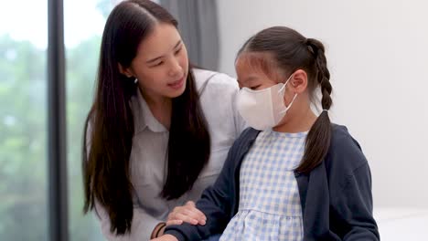 an asian mother gently reassures her masked daughter in a bright, modern room. natural daylight, soft focus, and steady camera enhance the supportive, emotional mood