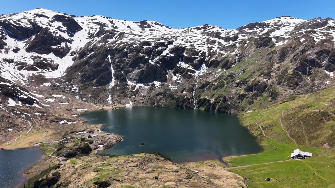 Scenic aerial view of Murgsee lake in Switzerland surrounded by snow capped alpine mountains, lush green meadows and clear blue sky in beautiful spring landscape, drone panning