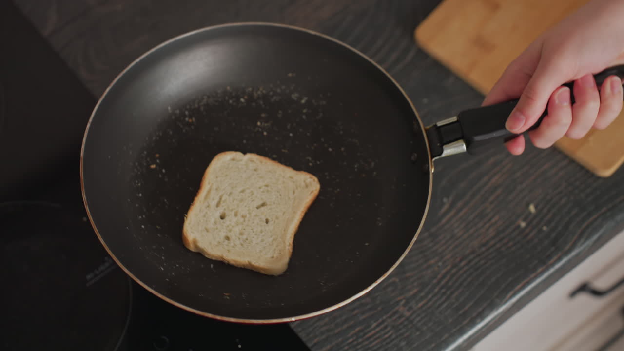 Close up of woman in green pajamas tossing bread slice upward inside frying pan above kitchen counter, motion blur and scattered crumbs