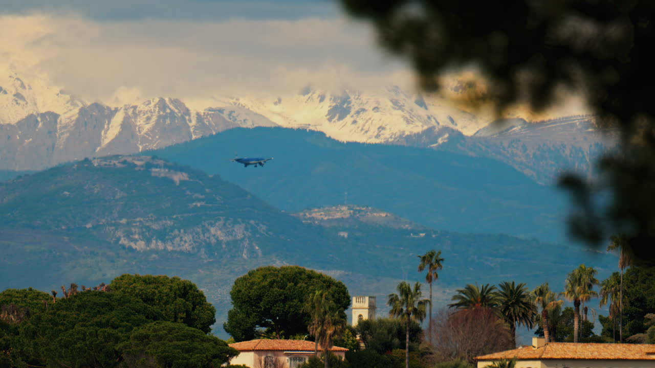 Distant view of an airplane flying above buildings surrounded by green trees with the mountains on the background on a cloudy day