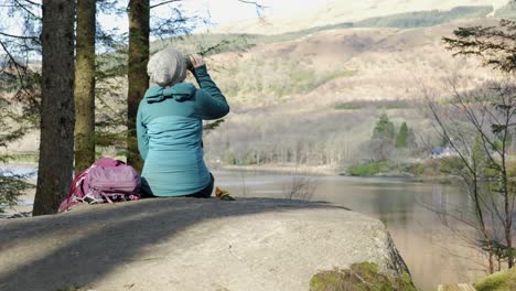 a woman sits on a rock eating her lunch and drinking tea while out for a walk in the forest, looking out across a scottish loch and hillside in the sun