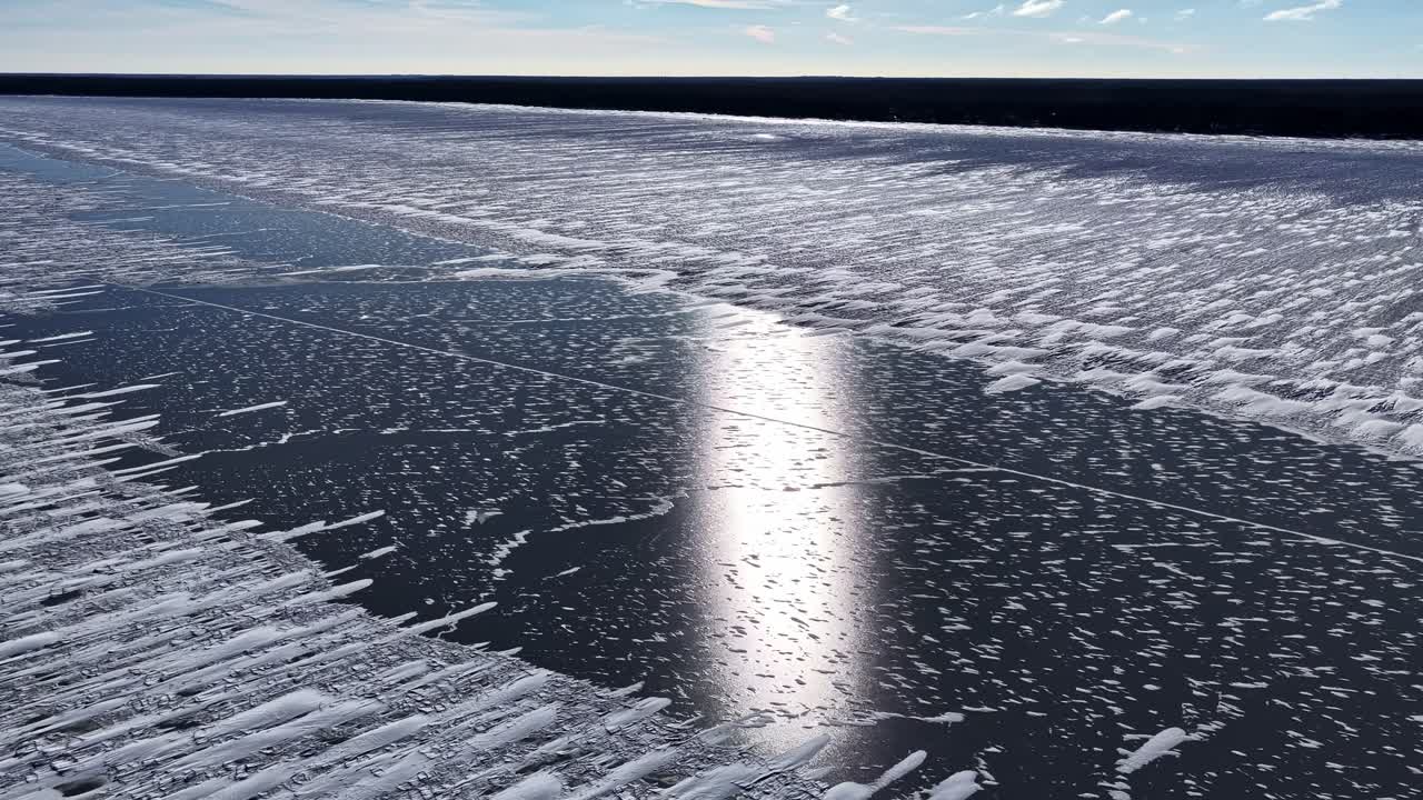 Tiefer Drohnenflug über einem zugefrorenen See mit windgeformten Schneemustern und heller Sonnenreflexion auf dunklem Eis, was eine idyllische Winterkulisse schafft.