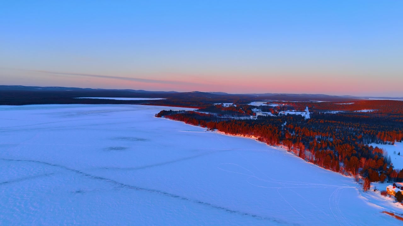 Vintersolnedgang over frossen innsjø og furuskog, Lappland. Luftfoto av rødt sollys som belyser snødekte trær, trehytter og islagt vidde i skumringen.