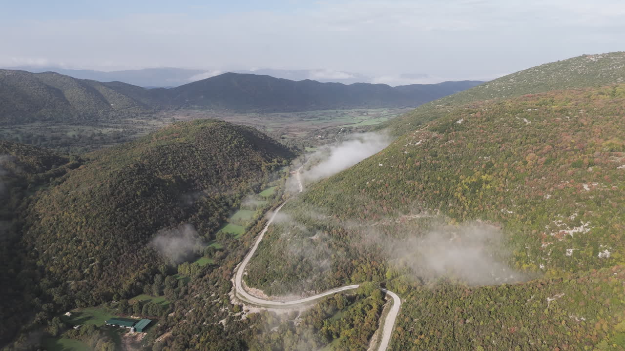 Aerial establishing view of a winding road in central Zagori, surrounded by misty landscapes and rugged terrain leading to Monodendri