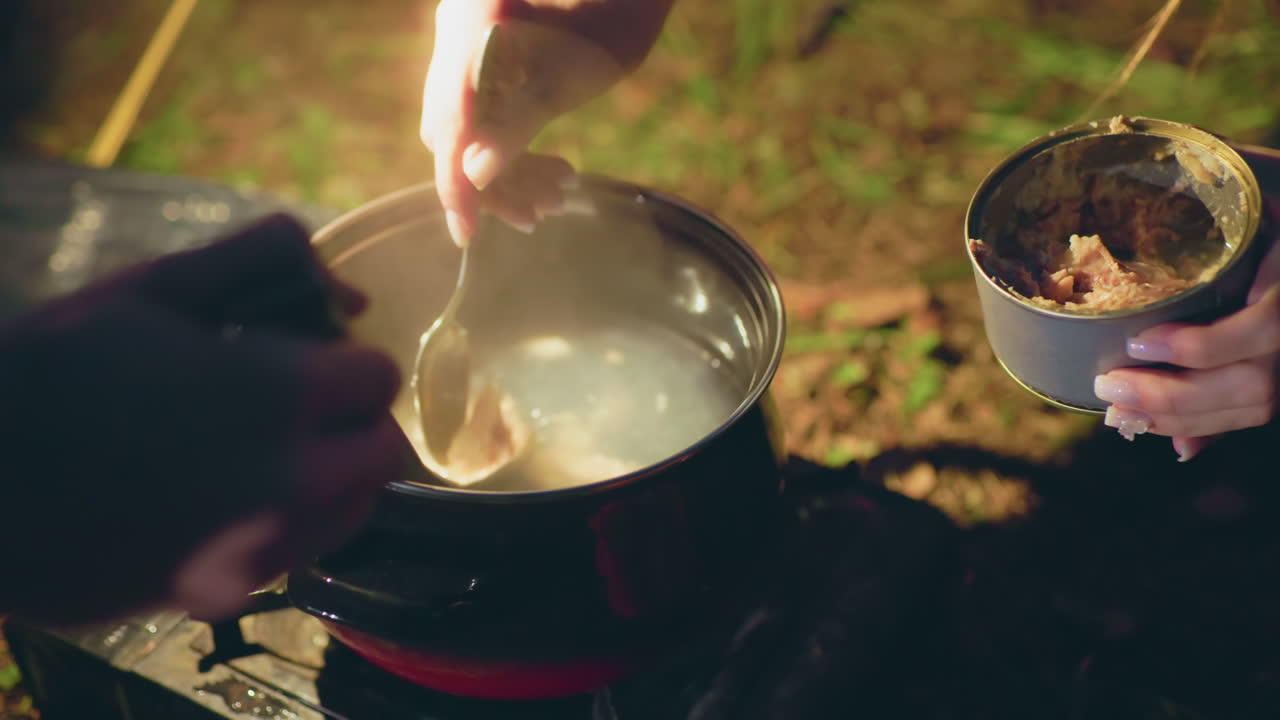 Close up of two people cooking outdoors at night as one stirs steaming pot on portable gas burner while other scoops canned food into boiling water, surrounded by forest ground and warm camping light