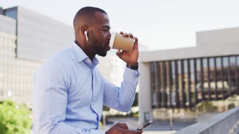 Hombre-Afroamericano-En-La-Ciudad-Parado-Al-Sol,-Tomando-Café,-Usando-Un-Teléfono-Inteligente