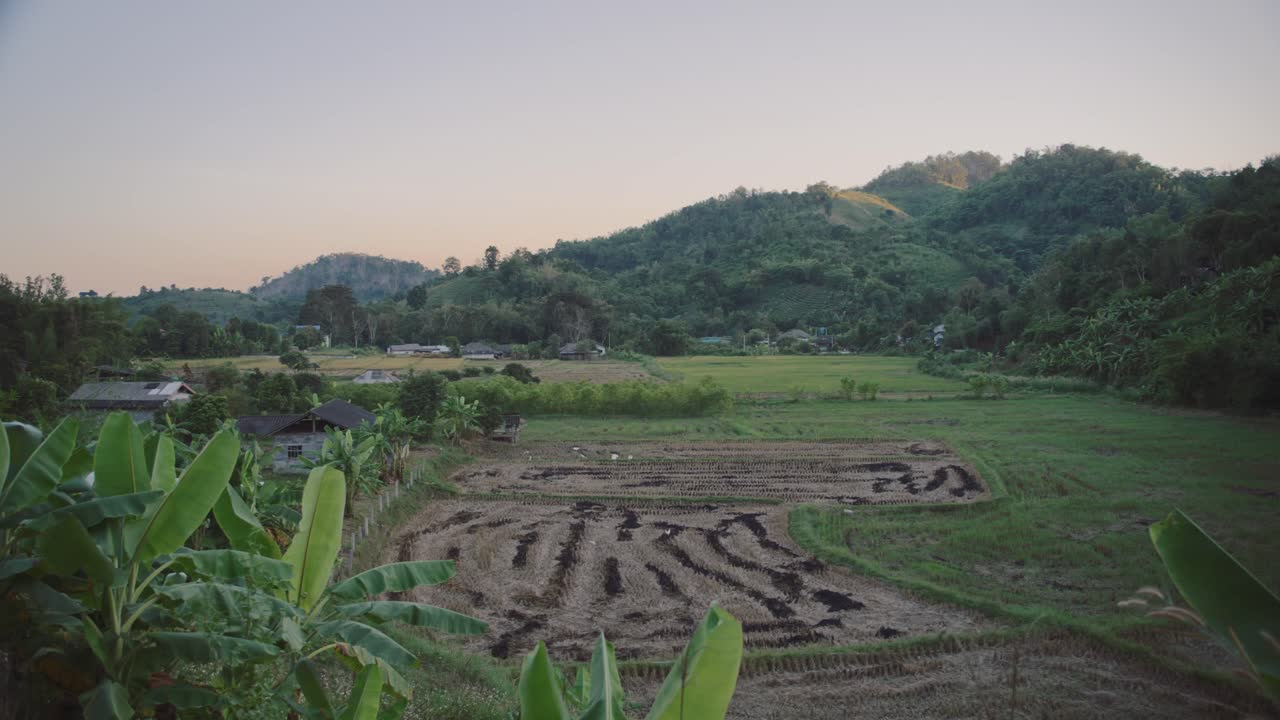 Panorama pedesaan yang menampilkan ladang padi hijau dan yang sudah dipanen dengan gunung-gunung di kejauhan dan rumah-rumah penduduk setempat