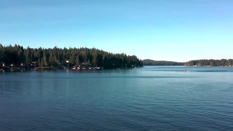 Aerial-dolly-push-in-above-placid-lake-as-speed-boats-rush-below,-homes-on-waters-edge-below-dense-pine-tree-forest