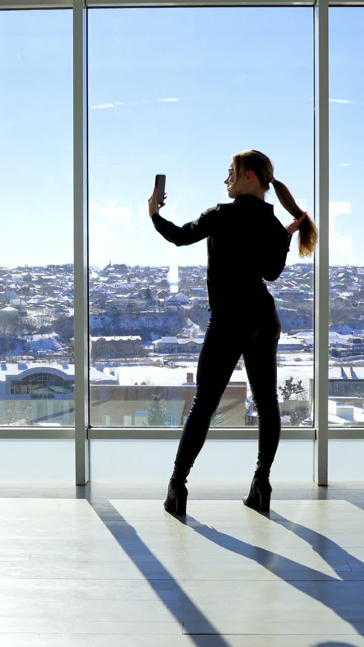Young woman taking selfie photo in the office. Beautiful girl standing by the large panoramic windows and taking photo of herself on a phone. Rear view. Vertical video