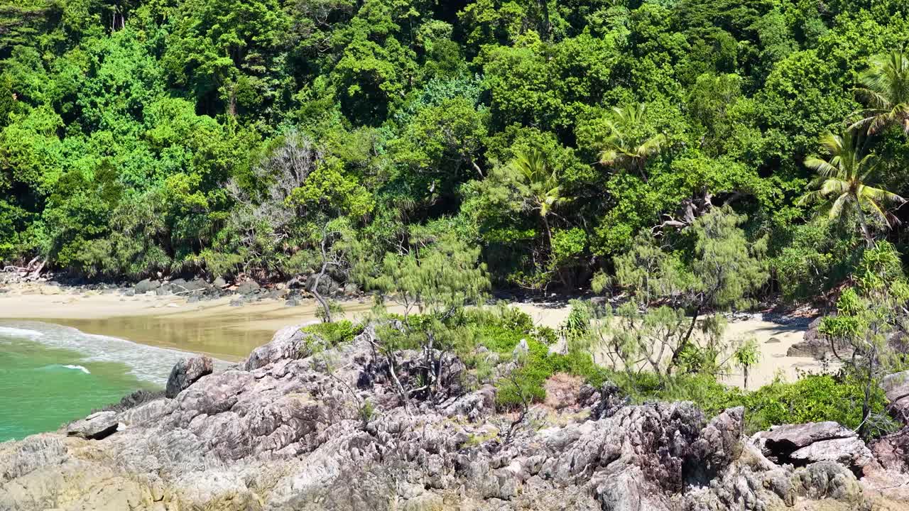 Drone camera rises above rocky shoreline, revealing dense tropical rainforest, sandy beach, and turquoise coastal waters in bright daylight