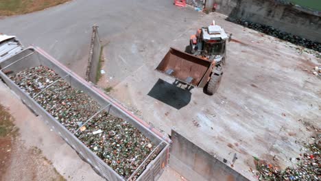 la excavadora de carretilla elevadora está agarrando vidrio y cargándolo en la parte trasera de un camión de transporte, la máquina de servicio pesado tiene un brazo largo equipado con un accesorio para agarrar vidrio, vista de drones, concepto de reciclaje de vidrio