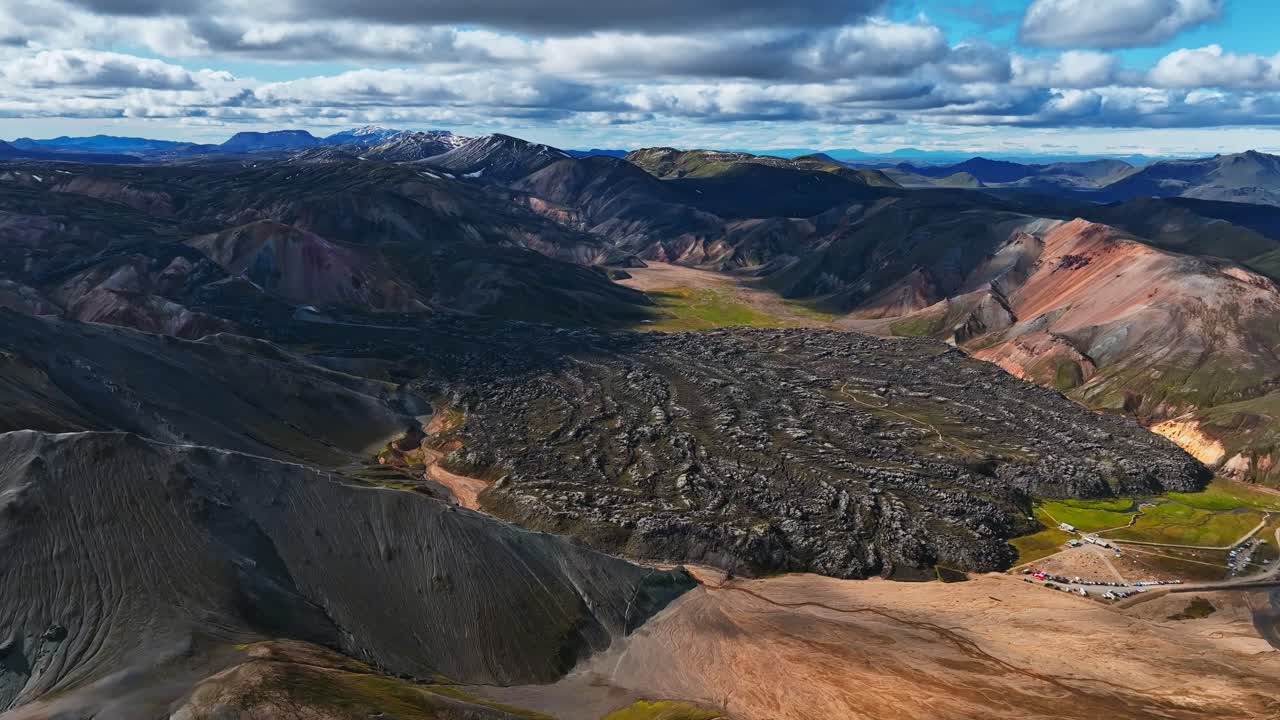 Aerial view of a rugged Icelandic lava field surrounded by colorful rhyolite mountains under a bright summer sky
